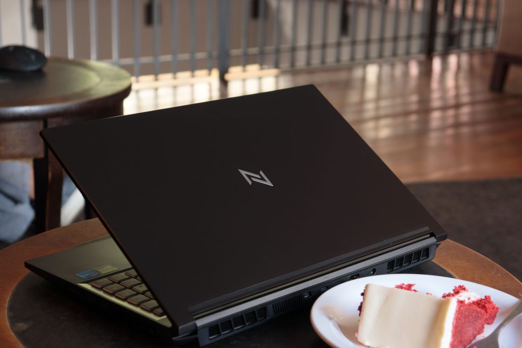 Notebook Gamer Neologic Journey fechado sobre uma mesa, ao lado de uma fatia de bolo, em ambiente interno iluminado.