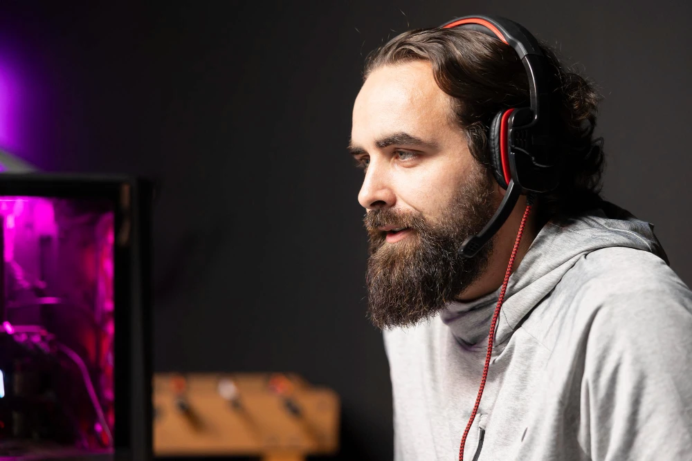 Homem com barba usando headset de jogos sentado na frente de um computador com iluminação rosa roxa, focado em jogar ou transmitir online.